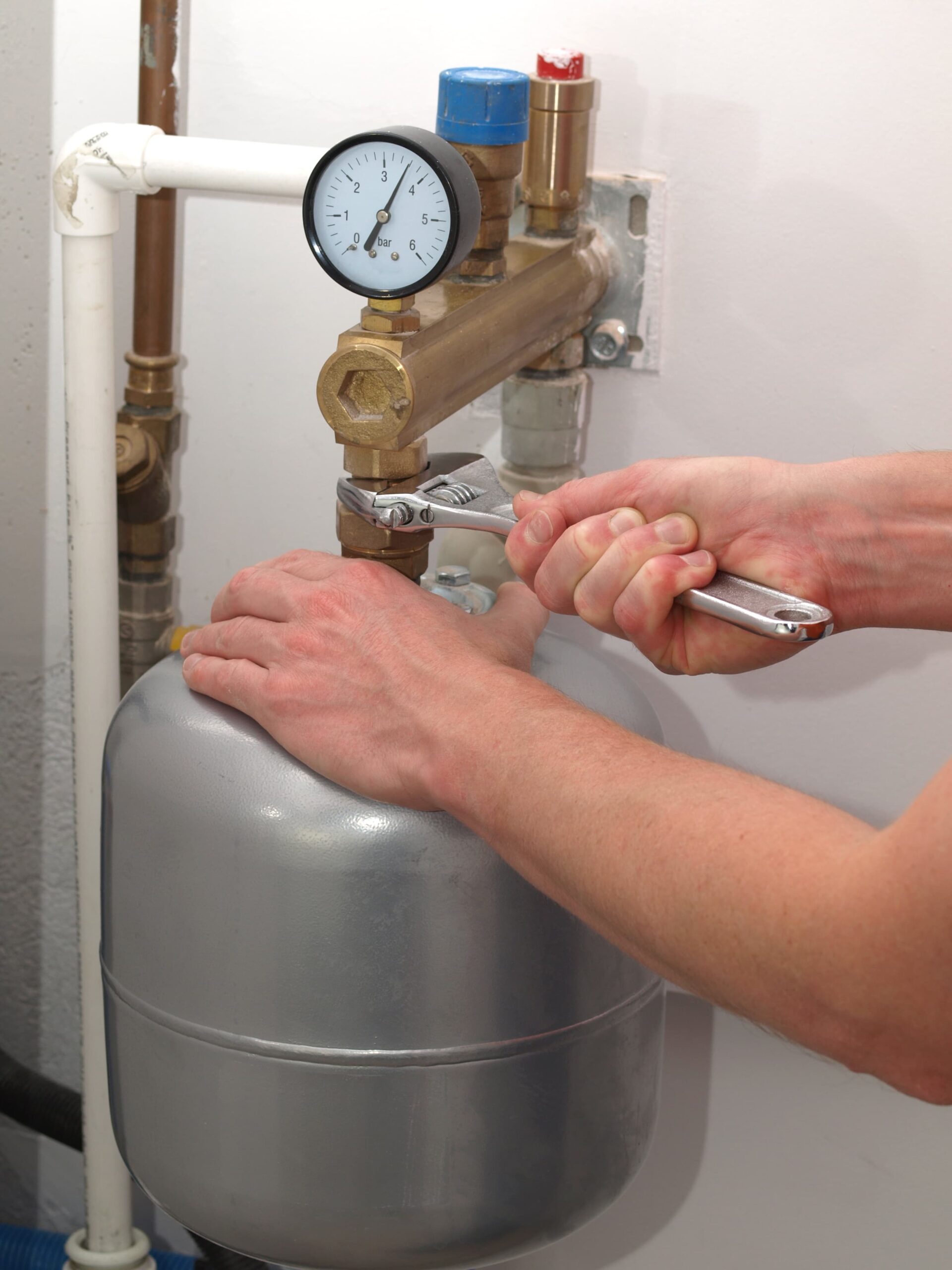 Person using a wrench to tighten a valve on a pressure tank in a plumbing system. The setup includes a pressure gauge and various brass and PVC pipes, indicating maintenance or adjustment of the system