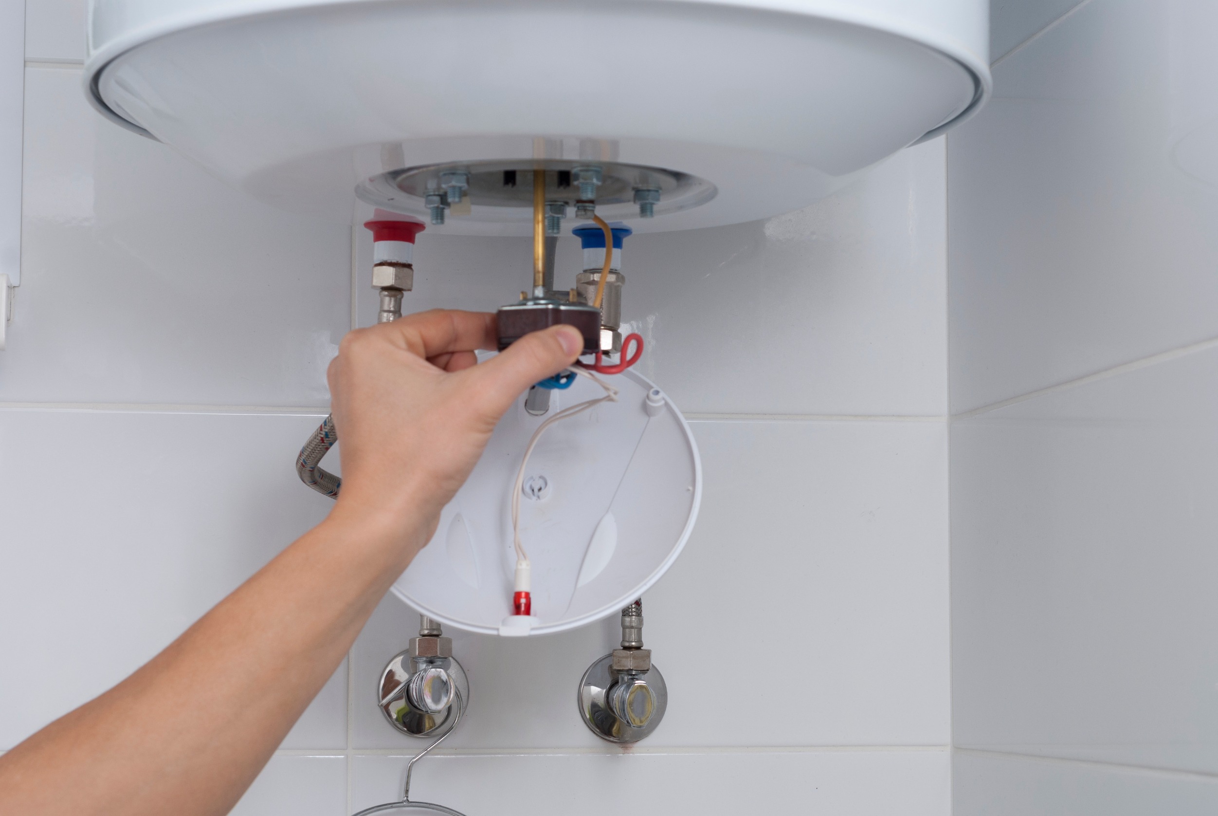 Person adjusting the settings on an electric water heater, with a focus on the internal wiring and plumbing connections, including hot and cold water valves and electrical components